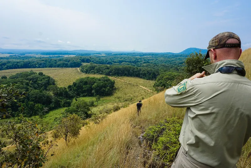 Un visiteur photographie un paysage de collines couvertes de savane et de bosquets dans la Lopé